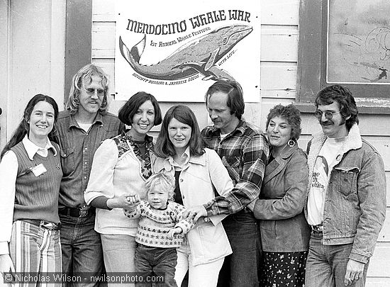 Organizers of the First Annual Mendocino Whale Festival pose outside Crown Hall with J.D. Mayhew's  poster, March 20, 1976. L to R: Ellen Findlay, Bill Wilson, Sue Golden, Brendan, Heidi and Barry Cusick, Sally and Lee Welty.